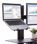 A woman stands at a desk using a black dual-monitor adjustable standing desk riser with a laptop on the top shelf and a keyboard on the lower shelf. The setup is shown against a clean white background.